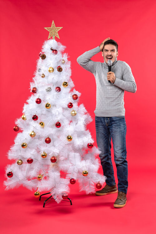 积极的家伙圣诞节气氛积极的家伙穿着牛仔裤站在装饰圣诞树附近 拿着麦克风 把手放在头上站着庆祝红色
