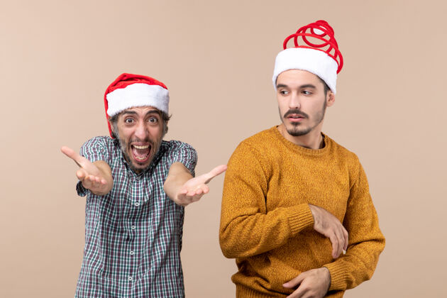 伙计们前视图两个戴着圣诞帽的家伙一个在叫喊 另一个在米色孤立背景下困惑两个家伙前面帽子