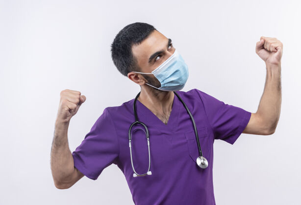 手势看着身边年轻的男医生穿着紫色手术服 戴着听诊器医用面罩 在孤立的白色背景上做着强烈的手势表情医生强壮