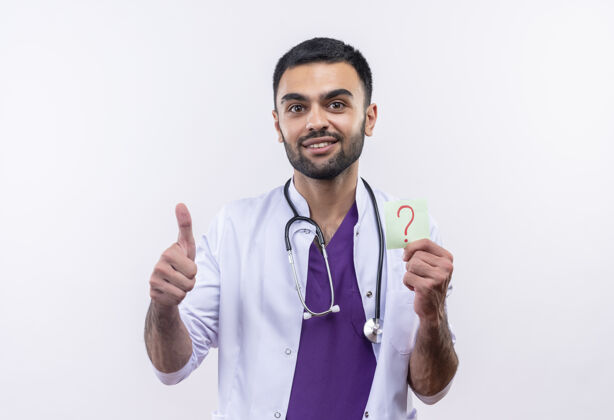 听诊器微笑着的年轻男医生穿着听诊器医用长袍拿着纸问号在孤立的白色背景上竖起大拇指白色问号男性
