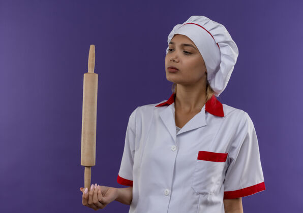 制服想着年轻的厨师 穿着厨师制服的女厨师在孤立的背景下看着手里的擀面杖厨师女性思考