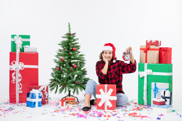 雪前视图年轻女子坐在圣诞礼物周围拿着白色墙上的时钟庆祝圣诞节快乐