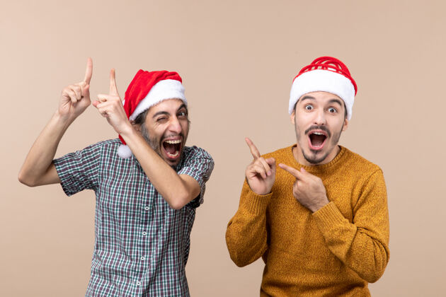 米色正面图两个戴着圣诞帽的兴高采烈的家伙 一个在米色孤立的背景上向另一个展示着什么兴高采烈肖像前面