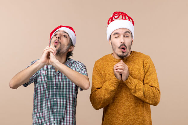 害怕前视图两个戴着圣诞帽的家伙一个在做手指枪的标志 另一个在米色的孤立背景上害怕两个男人帽子