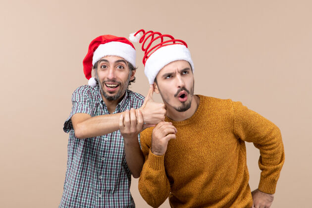 视图正面图两个戴着圣诞帽的家伙 一个微笑着 向另一个竖起大拇指 背景是米色的米色向上微笑