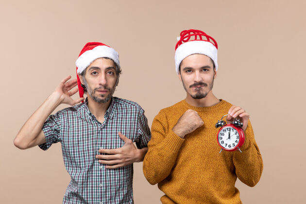 圣诞节前视图两个圣诞老人一只手放在胸前 另一只手拿着闹钟 背景是孤立的头饰男人两个圣诞男人