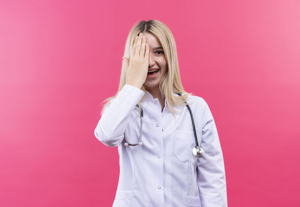 粉红色微笑的医生年轻的金发女孩穿着医用长袍戴着听诊器和牙套在孤立的粉红色背景上覆盖着眼睛支架金发牙科