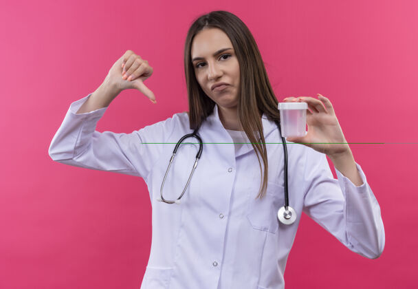 粉色悲伤的年轻医生女孩穿着听诊器医用长袍拿着空的大拇指趴在孤立的粉色背景上悲伤按住年轻