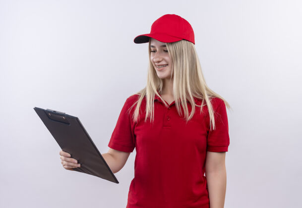 年轻带着微笑的小女孩穿着红色t恤 戴着帽子 戴着牙套 在孤立的白色背景下看着手边的剪贴板支架穿牙科