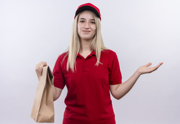 微笑微笑的递送年轻女孩穿着红色t恤和帽子拿着纸口袋在孤立的白色背景年轻白色穿着
