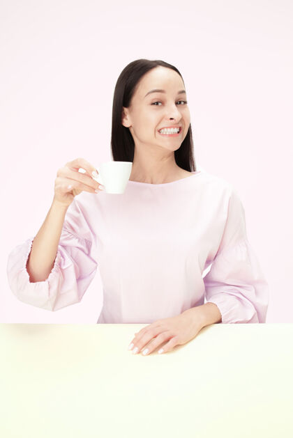 简约美丽的微笑的女人坐在粉红色的工作室里 手里拿着一杯咖啡 看起来很开心咖啡茶沉思