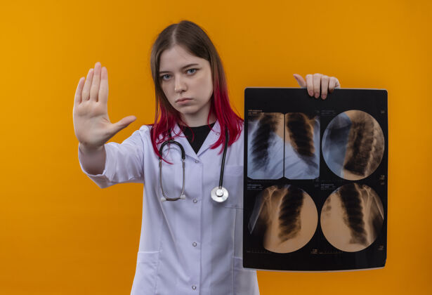 展示严格的年轻医生女孩穿着听诊器医用长袍拿着x光显示停止手势孤立的橙色背景长袍女孩穿着