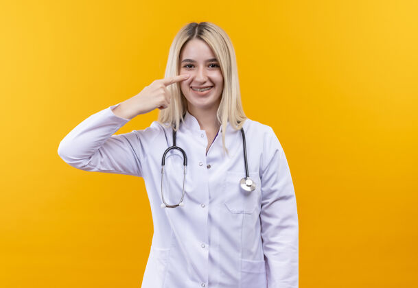 背带微笑的医生穿着医用长袍戴着牙套的听诊器的年轻女孩指着孤立的黄色背景上的眼睛医生穿着眼睛