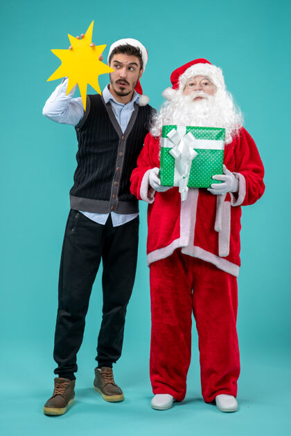 黄色前视图圣诞老人和年轻的男性谁持有黄色标志蓝色背景微笑圣诞老人服装
