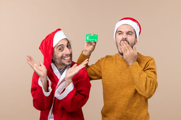 前面前视图两个圣诞节的家伙 一个穿着圣诞老人外套 另一个打着信用卡哈欠 背景是米色的圣诞老人圣诞节卡片