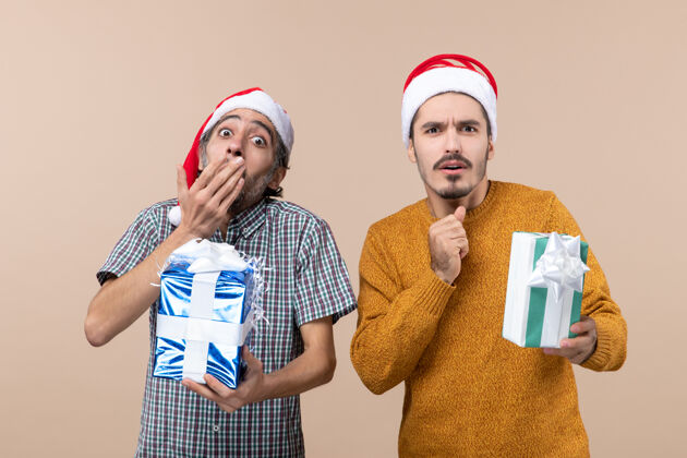 男人正面图两个戴着圣诞帽拿着礼物的好奇的家伙 一只手放在嘴上 背景是米色的伙计们成人礼物