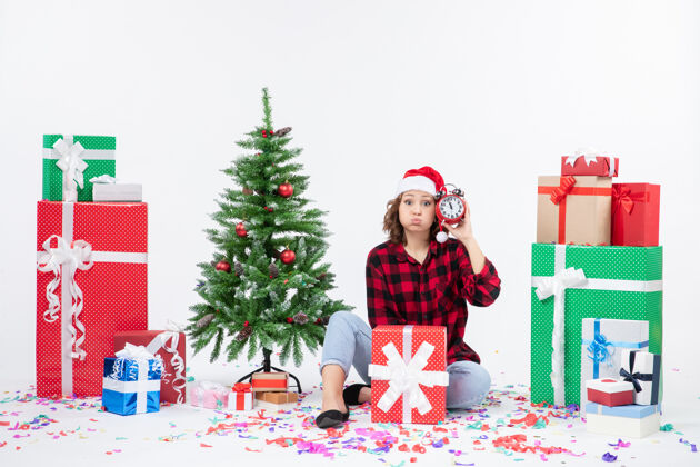 圣诞树前视图的年轻女子坐在圣诞礼物周围拿着白色墙上的时钟时钟女人节日