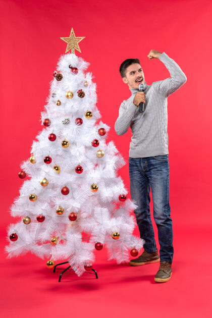圣诞圣诞节气氛积极的家伙穿着牛仔裤站在装饰圣诞树附近 手持麦克风展示他的力量权力积极的麦克风