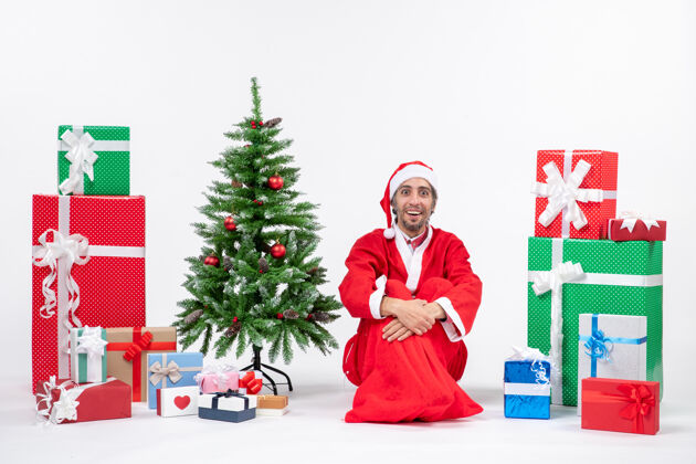 礼物微笑着惊讶的年轻人打扮成圣诞老人和礼物装饰圣诞树坐在地上的白色背景装饰微笑男人