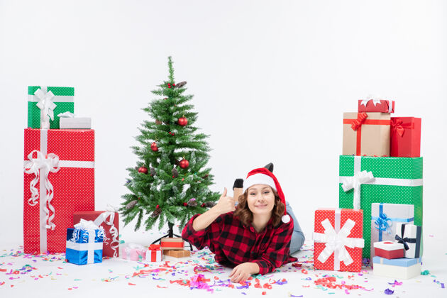 快乐年轻女子躺在白色墙壁上的圣诞礼物和小圣诞树周围的正视图圣诞树礼物装饰