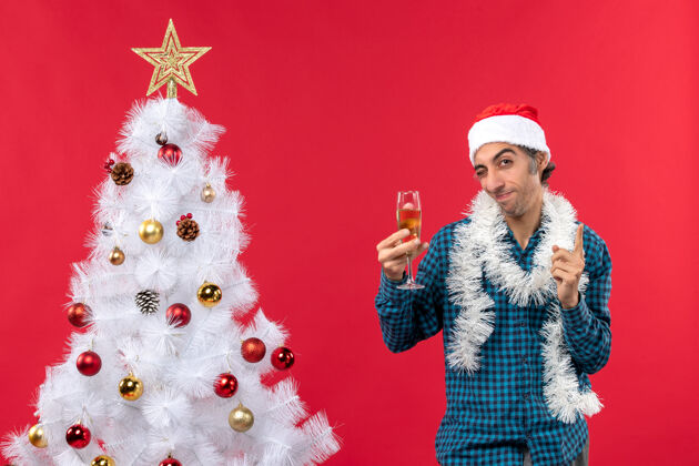 红色圣诞心情快乐疯狂情绪化的年轻人戴着圣诞老人的帽子 穿着蓝色条纹衬衫 在圣诞树旁举起一杯葡萄酒圣诞帽圣诞老人心情