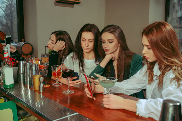 年轻女朋友在酒吧喝酒她们坐在一张摆着鸡尾酒的木桌旁她们穿着休闲服网络饮料干燥
