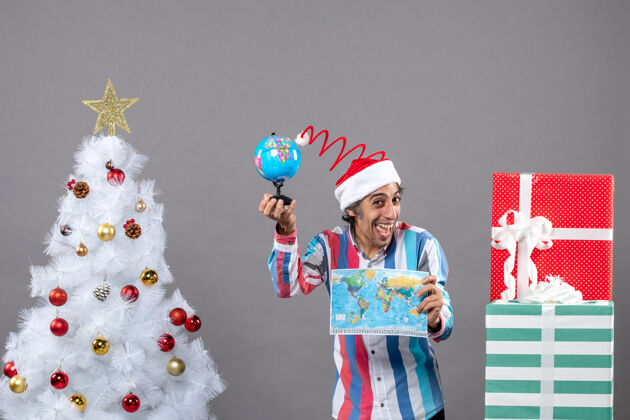 快乐前视图欣喜若狂的男子拿着世界地图和地球仪在圣诞树旁送礼物旅行微笑人