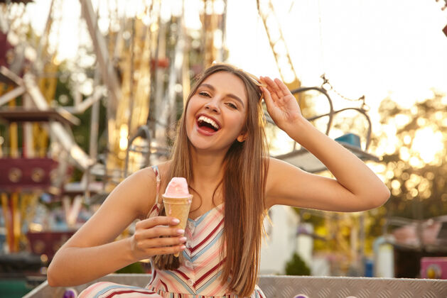 微笑在户外拍摄的美丽的年轻快乐的女性长发吃冰淇淋锥 而通过游乐园散步 笑得很开心 张大嘴巴 摘下墨镜宽可爱温暖