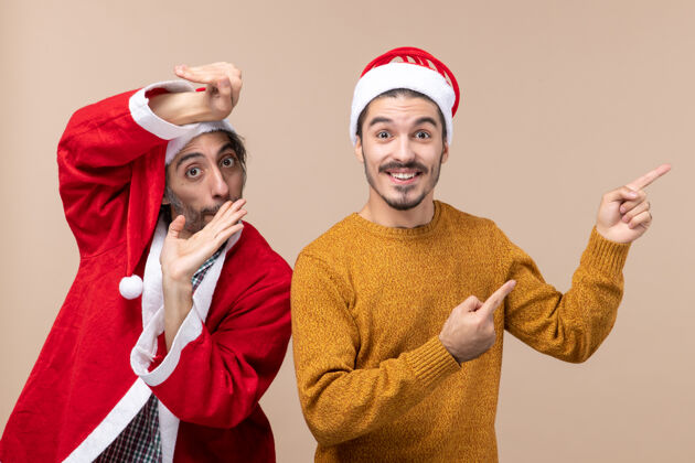 前面正面图两个戴着圣诞帽的朋友 一个在米色孤立背景上用快乐的脸显示方向米色微笑肖像