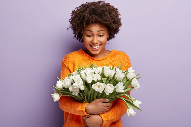 满意温柔积极的非洲裔美国女士拥抱着温柔的白色花朵 微笑着看着郁金香 穿着橙色的套头衫非洲女性种族