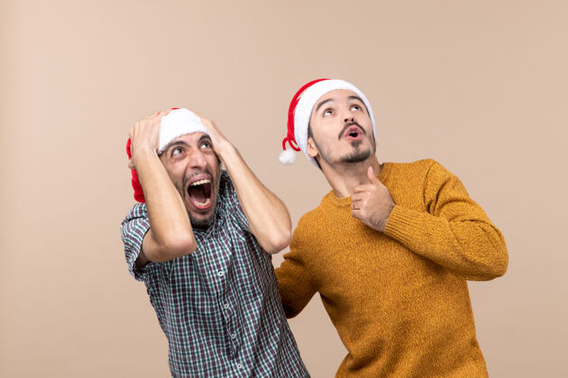 年轻前视图两个戴着圣诞帽的吓坏了的家伙在米色孤立的背景下看着高处前面表情伙计们