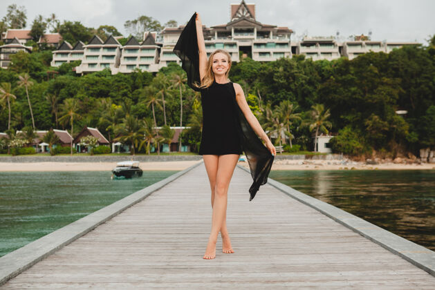 女士穿着黑色连衣裙的美丽迷人的女人在豪华度假酒店的码头上摆姿势 暑假 热带海滩头发挥舞奢华