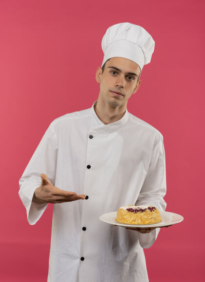 穿年轻的男厨师穿着厨师制服 拿着手指头 把蛋糕放在盘子上孤立的粉红色墙壁上拿盘子年轻