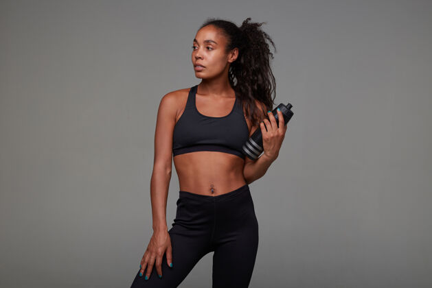 女士迷人的年轻黑皮肤女运动员卷曲的长发手持健身瓶 站在黑色运动上衣和紧身裤健身摆姿势卷发