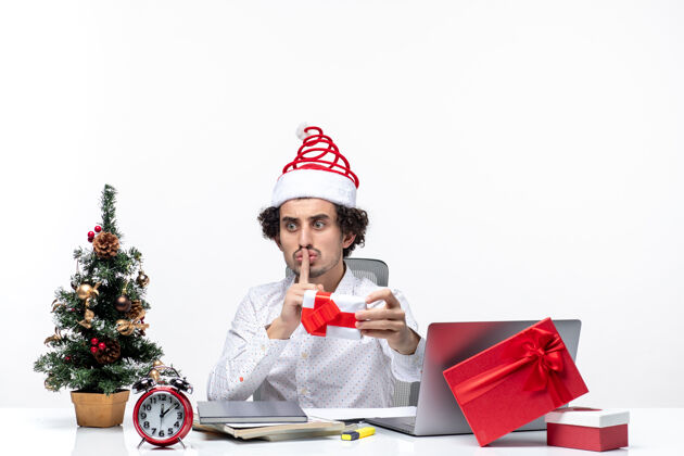 东西圣诞节心情惊讶的商人拿着圣诞老人的帽子拿着他的礼物 做着沉默的手势看着白色背景上的东西成人帽子手势