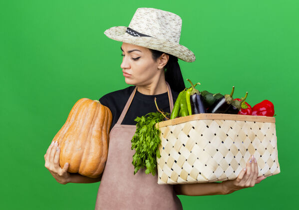 帽子年轻漂亮的女园丁围着围裙 戴着帽子 手里拿着装满蔬菜和南瓜的箱子 严肃地看着它抱着南瓜看