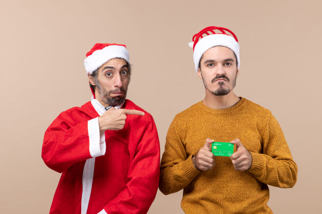 信用卡前视图两个快乐的家伙 一个穿着圣诞老人外套 手指尖 另一个在米色背景下 有信用证前面男人外套