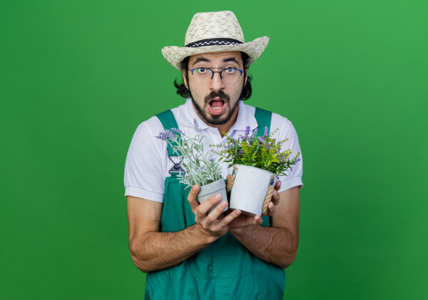 男人年轻的留着胡须的园丁穿着连体衣戴着帽子拿着盆栽植物感到惊讶惊讶园丁连身衣