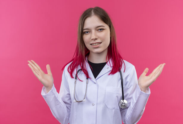 粉红色带着微笑的年轻医生女孩穿着听诊器医用长袍 在粉色孤立的背景上展开双手传播长袍手