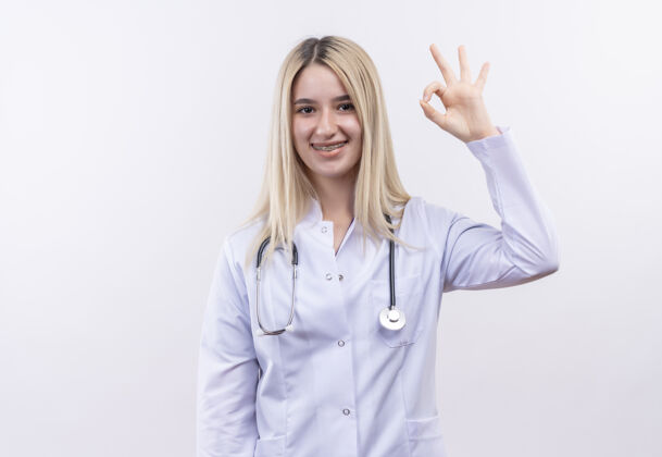 年轻微笑的医生年轻的金发女孩戴着听诊器和医用长袍 戴着牙套 在孤立的白色背景上显示出良好的姿势长袍医生展示