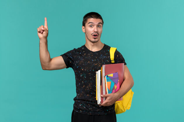 学生前视图中的男学生穿着深色t恤 背着黄色背包 手里拿着抄写本和文件 手指举在蓝色的墙上男性人黑暗