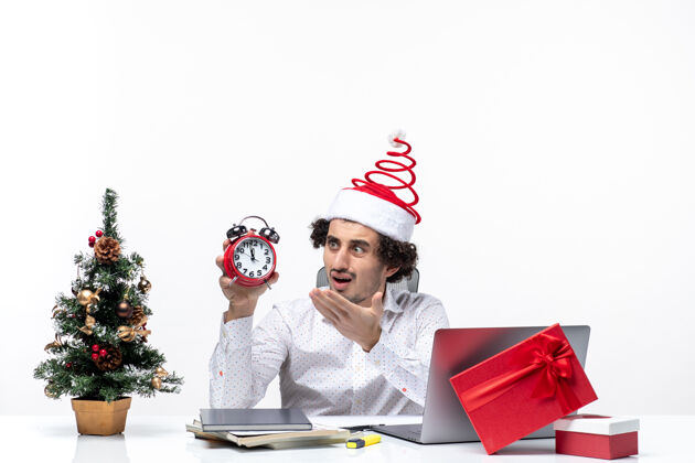 圣诞老人年轻关切的商人惊讶地戴着圣诞老人的帽子 拿着白色背景的办公室里的钟看着礼物微笑办公室