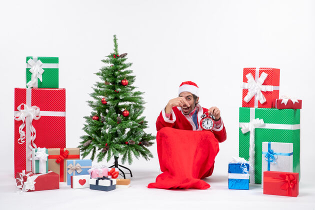 礼物微笑的年轻人坐在地上 拿着时钟 指着旁边的礼物和装饰圣诞树庆祝新年或圣诞节年轻人礼物圣诞树