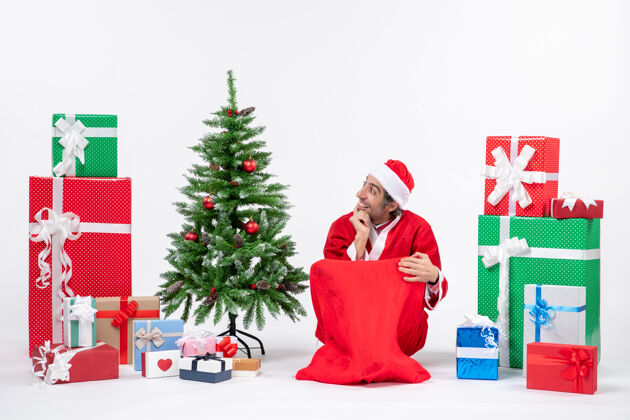 右边笑容满面的年轻人打扮成圣诞老人 拿着礼物和装饰好的圣诞树坐在地上 专注于白色背景右侧的某样东西圣诞树礼物男人