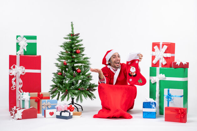 坐着微笑着积极的圣诞老人坐在地上 拿着圣诞袜靠近礼物和白色背景装饰的新年树袜子圣诞老人地面