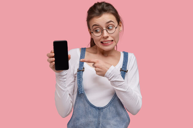 应用程序情绪化的年轻女子照片指着现代小玩意屏幕 犹豫是否购买智能手机 神情困惑技术眼镜手指