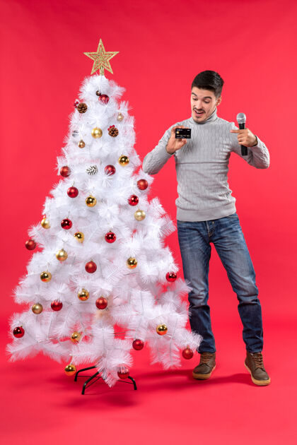 时尚圣诞节心情与情绪化的家伙站在装饰圣诞树附近 手持麦克风 指着他的手机快乐树心情