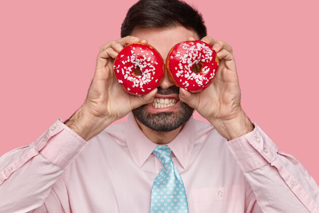 甜点特写镜头中 满脸胡须的男人咬紧牙关 用甜甜圈遮住眼睛 穿着正式 站在粉红色的墙上阳刚衣服学生