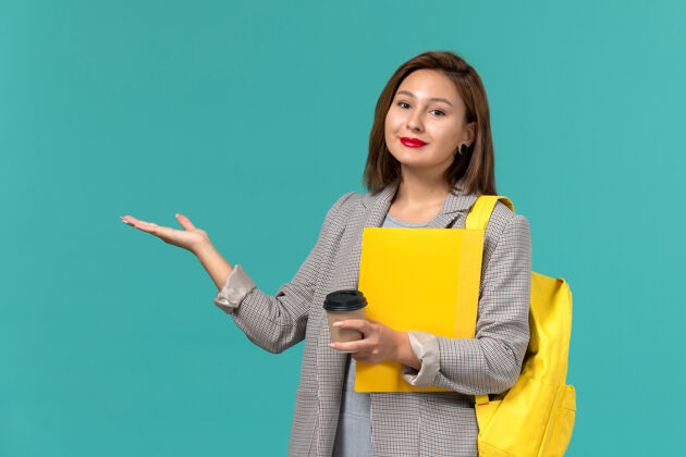 穿着身穿灰色夹克 背着黄色背包 在浅蓝色墙上拿着文件和咖啡的女学生的正视图人举行学校