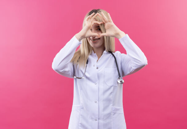 粉色微笑的医生年轻的金发女孩穿着医用长袍戴着听诊器和牙套在孤立的粉红色背景上显示心脏的姿态展示手势医学